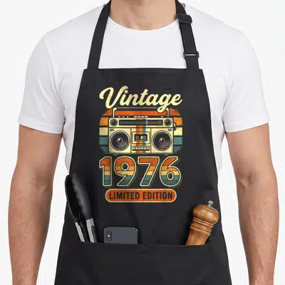 Tablier de cuisine noir avec illustration vintage d'une boombox et l'année 1976 en lettres colorées rayées.