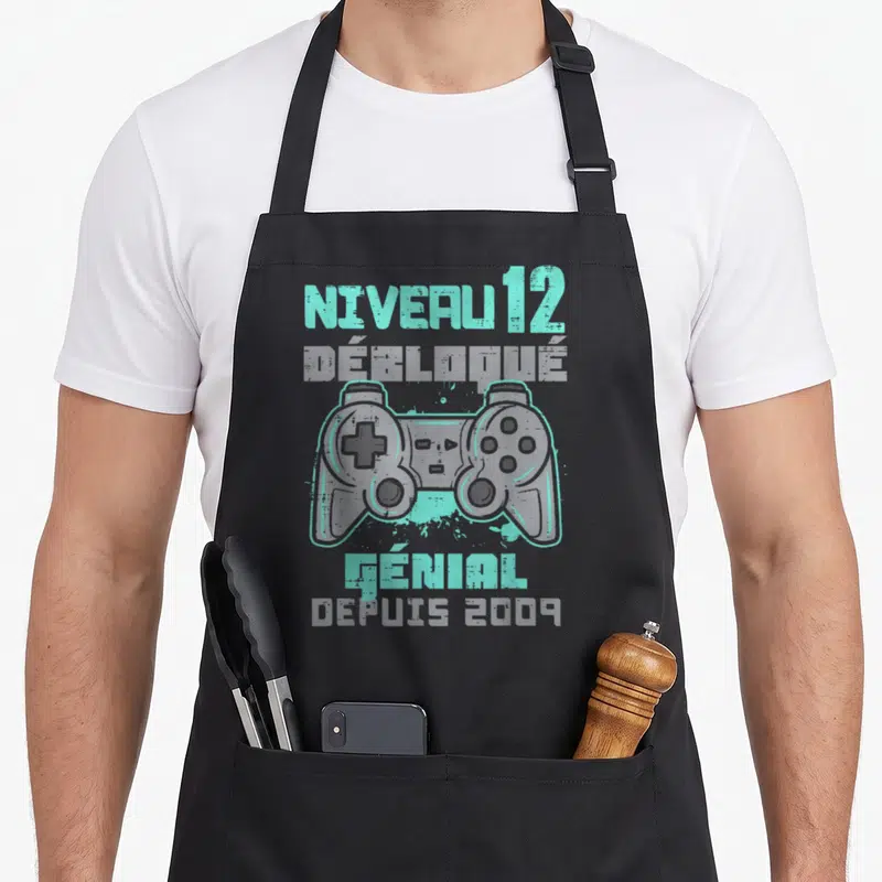 Tablier de cuisine noir avec inscription Niveau 12 Débloqué et illustration de manette de jeu vidéo gris et turquoise.