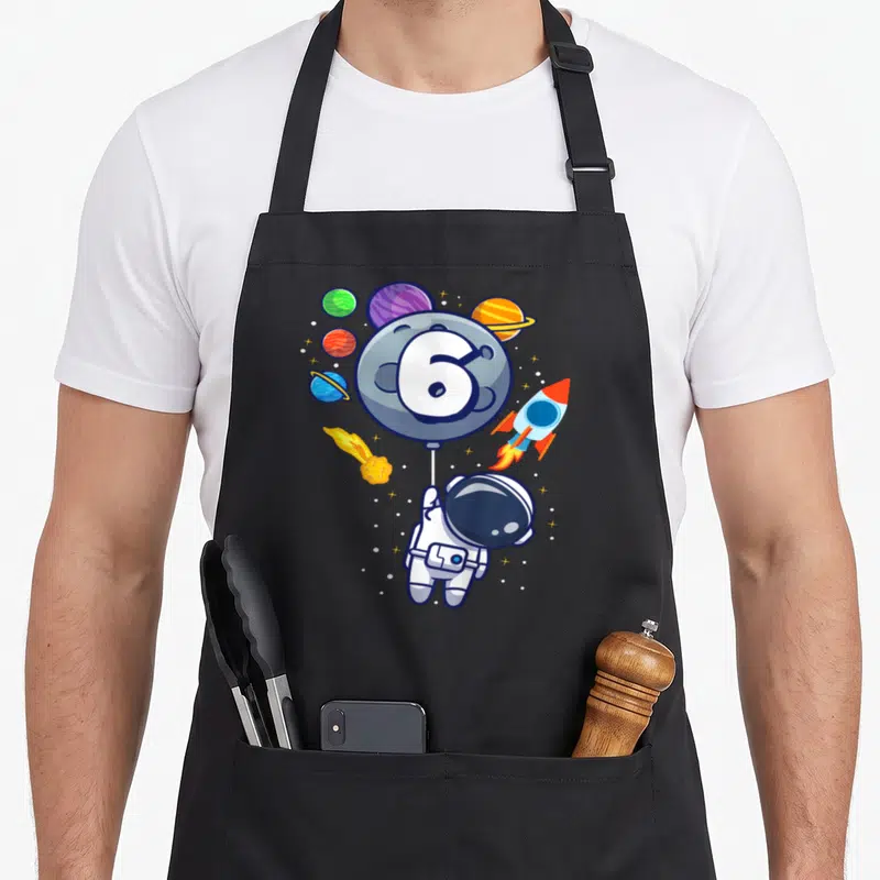 Tablier de cuisine noir avec astronaute tenant un ballon 6 entouré de planètes et d'une fusée.