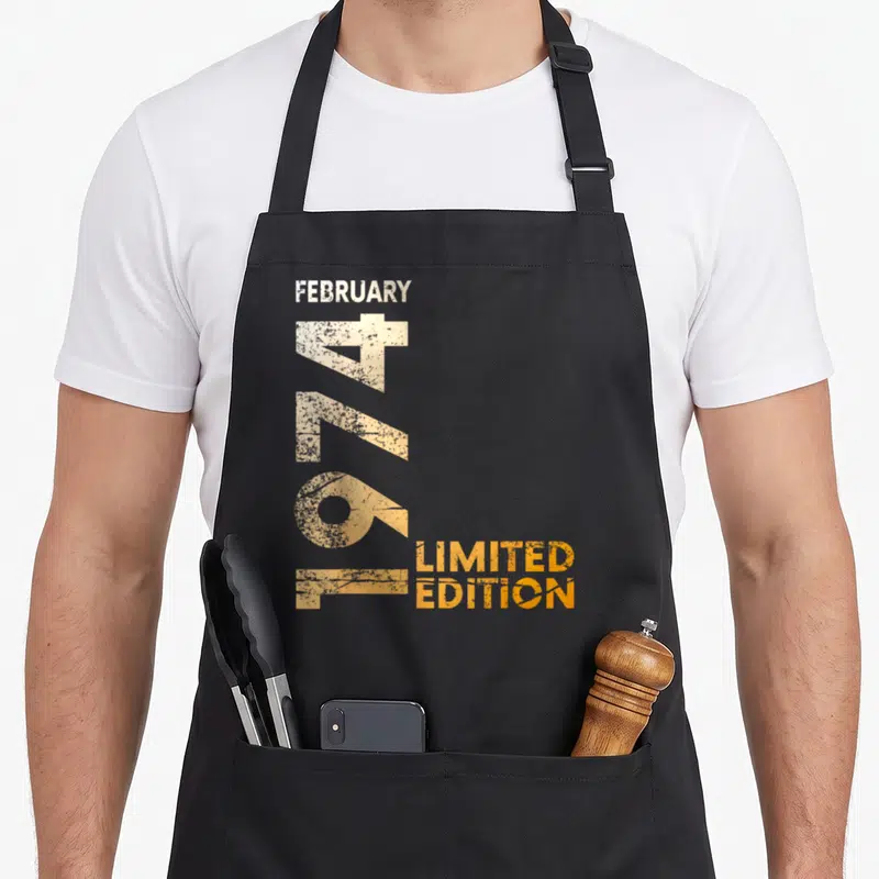 Tablier de cuisine noir avec l'inscription February 1974 Limited Edition en typographie vintage orange et beige sur le devant.