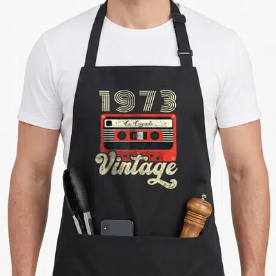 Tablier de cuisine noir avec illustration cassette rouge 1973 et texte Vintage La Légende en typographie rétro.
