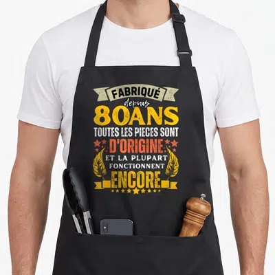 Tablier de cuisine noir avec inscription fabrique depuis 80 ans toutes les pieces sont dorigine et la plupart fonctionnent encore entouré d'étoiles et de plumes dorées.