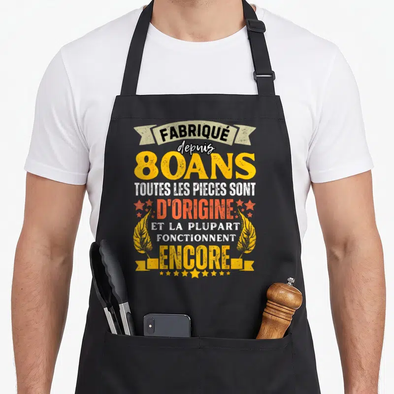 Tablier de cuisine noir avec inscription fabrique depuis 80 ans toutes les pieces sont d'origine et la plupart fonctionnent encore entouré d'étoiles et de plumes dorées.