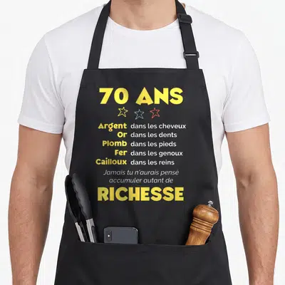 Tablier de cuisine noir avec inscription 70 ans Richesse et liste humoristique sur fond gris uni.