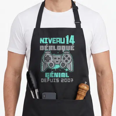 Tablier de cuisine noir avec illustration de manette de jeu vidéo et texte Niveau 14 Débloqué Génial Depuis 2007 en turquoise et gris.