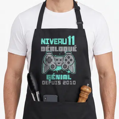 Tablier de cuisine noir avec illustration manette de jeu et texte Niveau 11 Débloqué Génial depuis 2010 en turquoise et gris.
