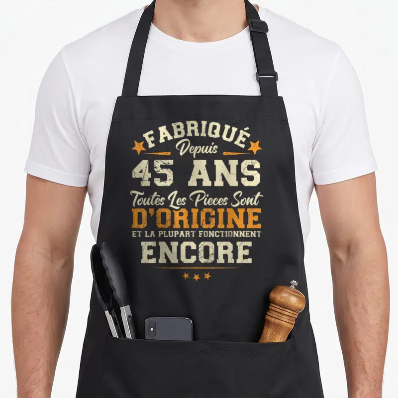Tablier de cuisine noir avec typographie vintage beige et orange proclamant être fabriqué depuis 45 ans avec des pièces d'origine encore fonctionnelles.