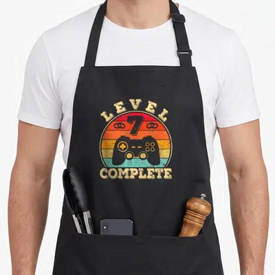 Tablier de cuisine noir avec motif rétro Level 7 Complete, manette de jeu vidéo et coucher de soleil coloré.