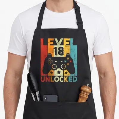 Tablier de cuisine noir avec illustration manette de jeu vidéo et texte Level 18 Unlocked en couleurs rétro.