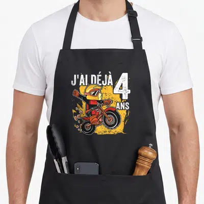 Tablier de cuisine noir avec un pilote de moto rouge et jaune levant le bras sous l'inscription J'ai déjà 4 ans.