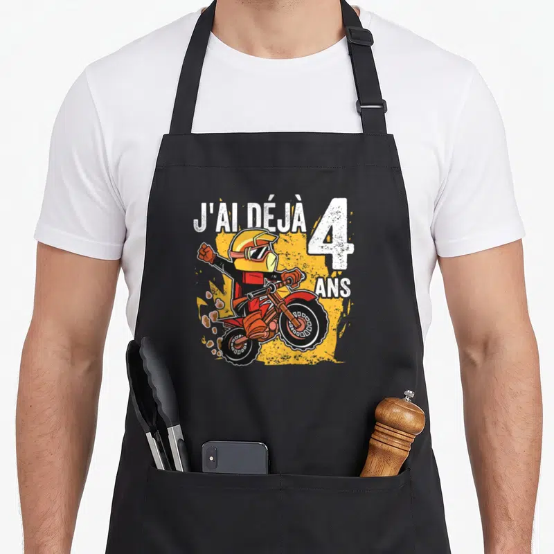 Tablier de cuisine noir avec un pilote de moto rouge et jaune levant le bras sous l'inscription J'ai déjà 4 ans.