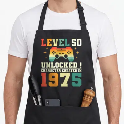Tablier de cuisine noir avec manette de jeu vidéo rétro colorée et texte Level 50 Unlocked 1975 imprimé au centre.