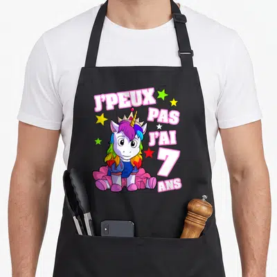 Tablier de cuisine noir avec licorne rose et texte J'peux pas j'ai 7 ans entouré d'étoiles colorées.