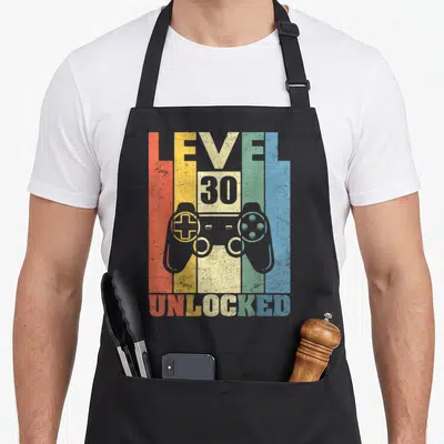 Tablier de cuisine noir avec illustration manette vidéo et texte Level 30 Unlocked multicolore style vintage.