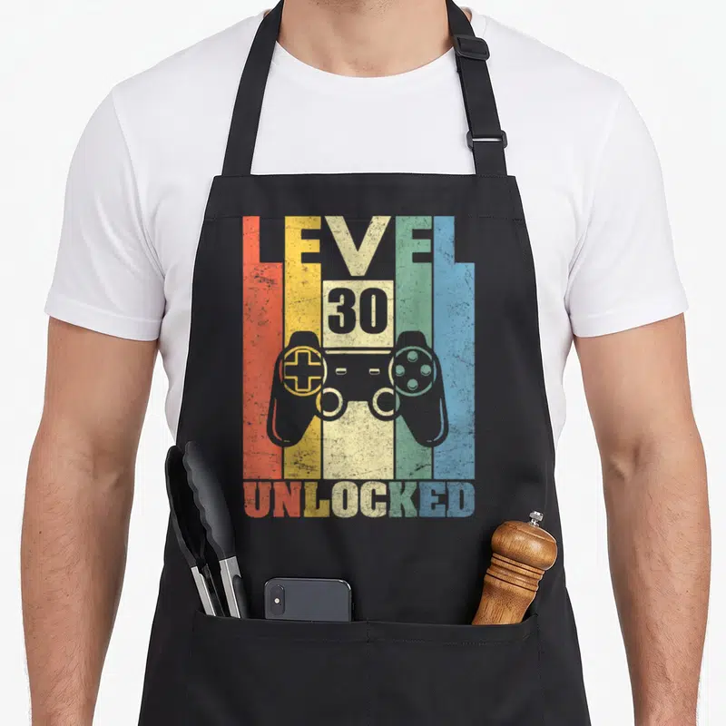 Tablier de cuisine noir avec illustration manette vidéo et texte Level 30 Unlocked multicolore style vintage.