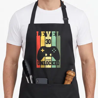 Tablier de cuisine noir avec illustration manette de jeu vidéo et texte Level 30 débloqué sur fond de barres colorées verticales.
