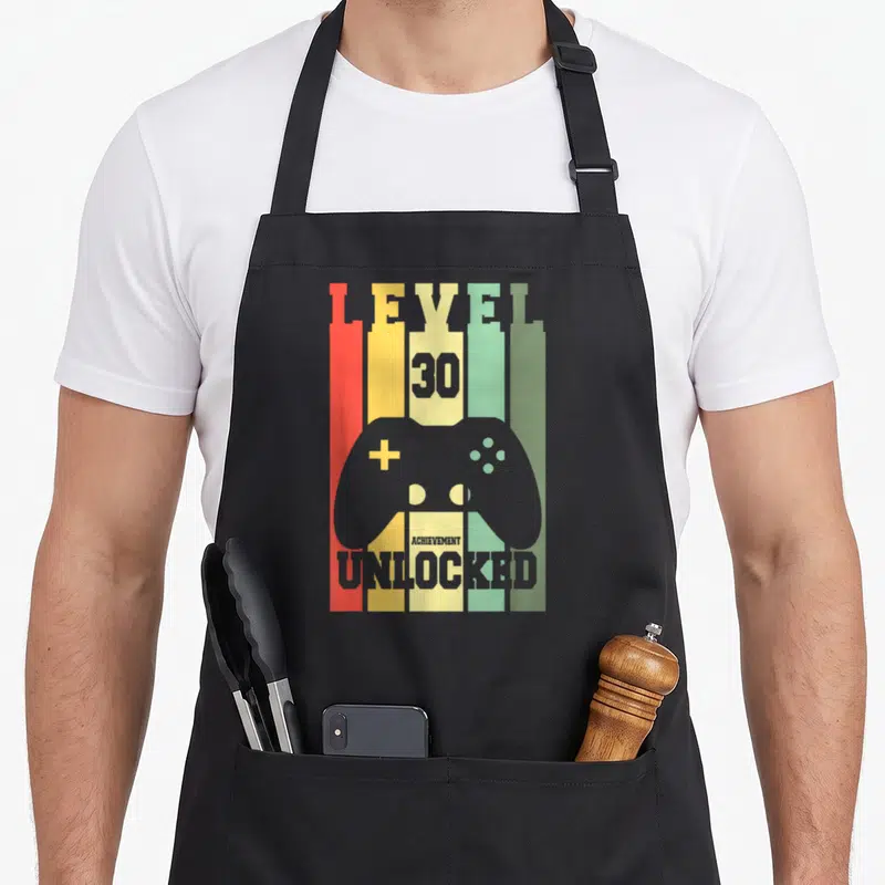 Tablier de cuisine noir avec illustration manette de jeu vidéo et texte Level 30 débloqué sur fond de barres colorées verticales.