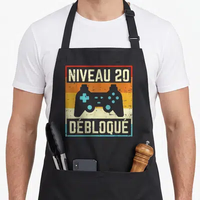 Tablier de cuisine noir avec illustration manette vidéo et texte Niveau 20 Débloqué sur fond coloré rétro.