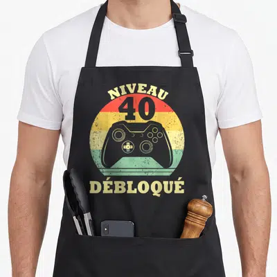 Tablier de cuisine noir avec illustration manette de jeu vidéo et texte Niveau 40 Débloqué sur fond coloré circulaire.