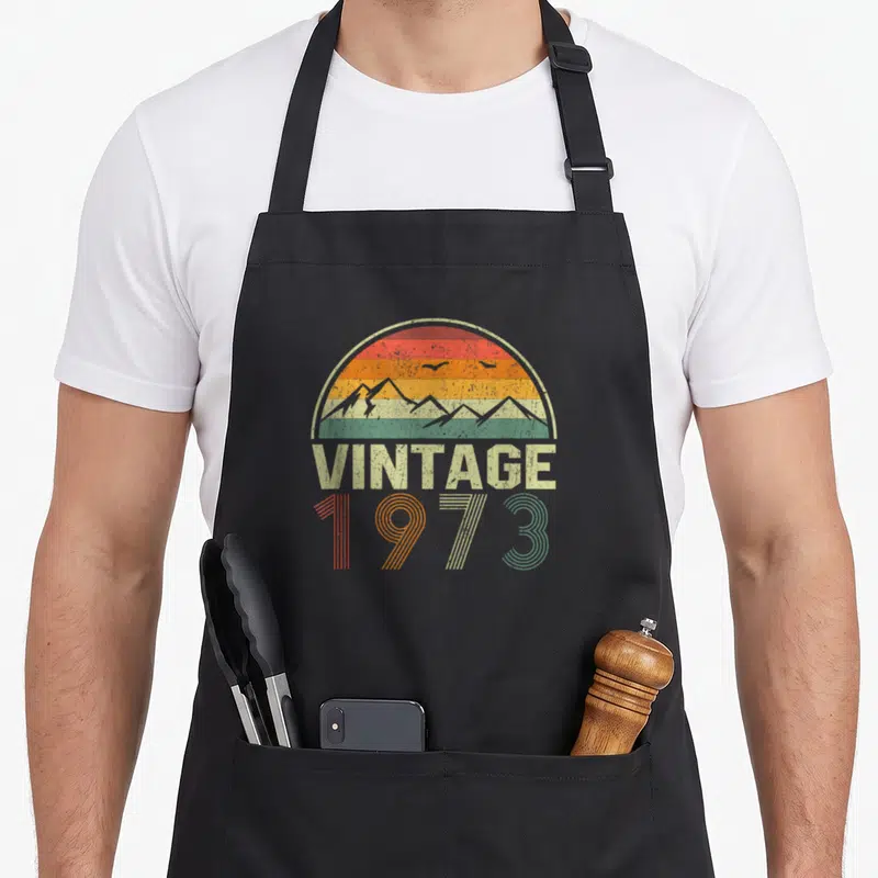 Tablier de cuisine noir avec motif vintage 1973 soleil couchant et montagnes en couleurs dégradées.