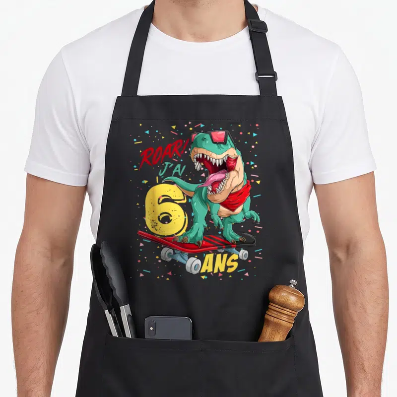 Tablier de cuisine noir avec dinosaure vert sur planche à roulettes, texte Roar J'ai 6 ans et confettis multicolores.