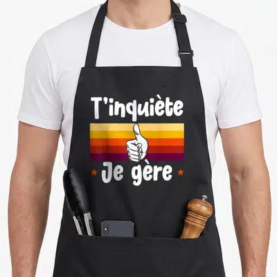 Tablier de cuisine noir avec inscription T'inquiète Je gère et illustration de main faisant un pouce levé sur fond coloré.