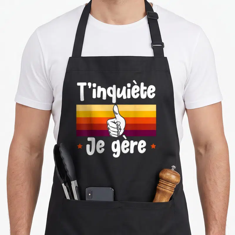 Tablier de cuisine noir avec inscription T'inquiète Je gère et illustration de main faisant un pouce levé sur fond coloré.
