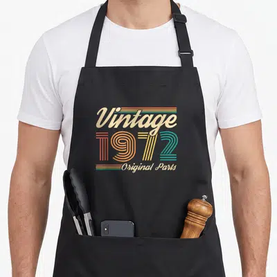 Tablier de cuisine noir avec typographie vintage 1972 Original Parts multicolore sur fond uni.