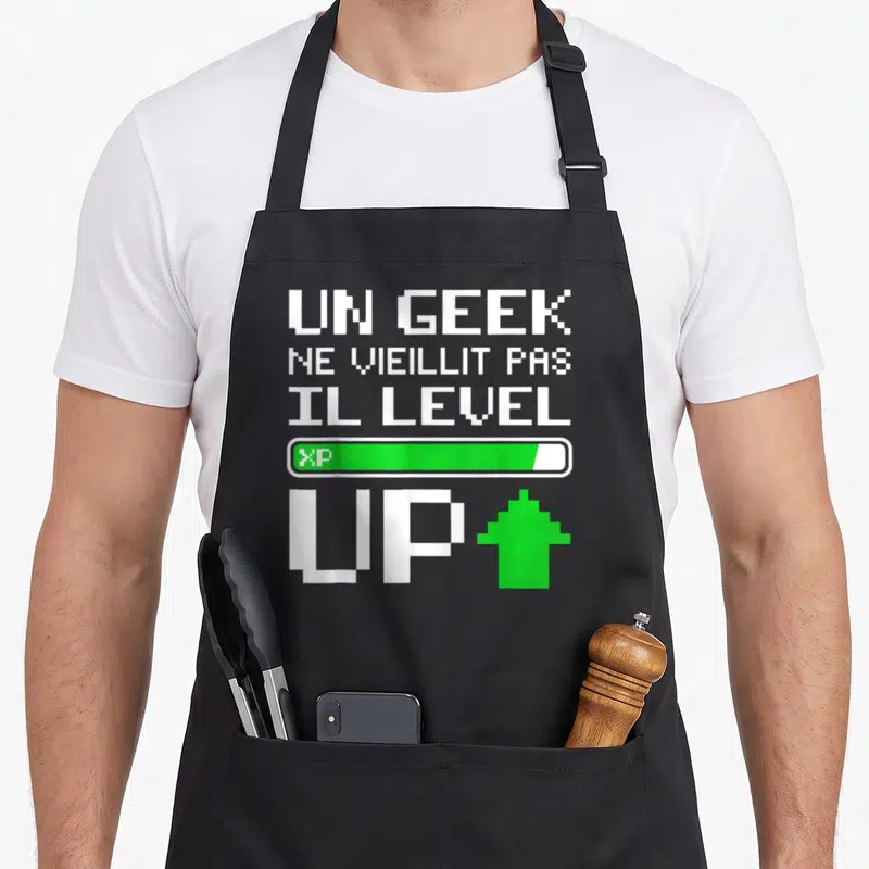 Tablier de cuisine noir avec texte pixelisé blanc Un Geek Ne Vieillit Pas Il Level Up et barre d'expérience verte.