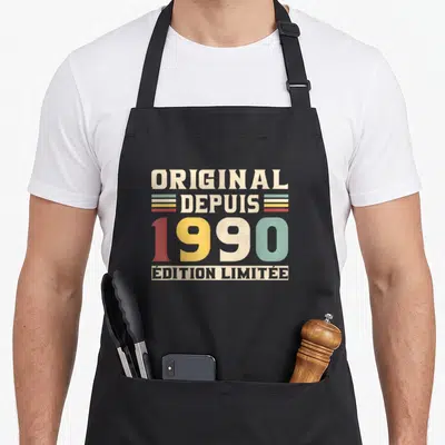 Tablier de cuisine noir avec typographie vintage colorée indiquant l'année de naissance 1990 et des bandes décoratives rétro.
