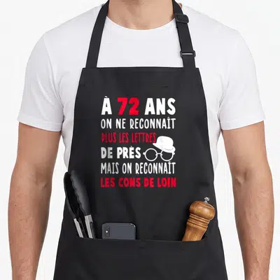 Tablier de cuisine noir avec texte blanc et rouge à 72 ans et illustration de lunettes et chapeau.
