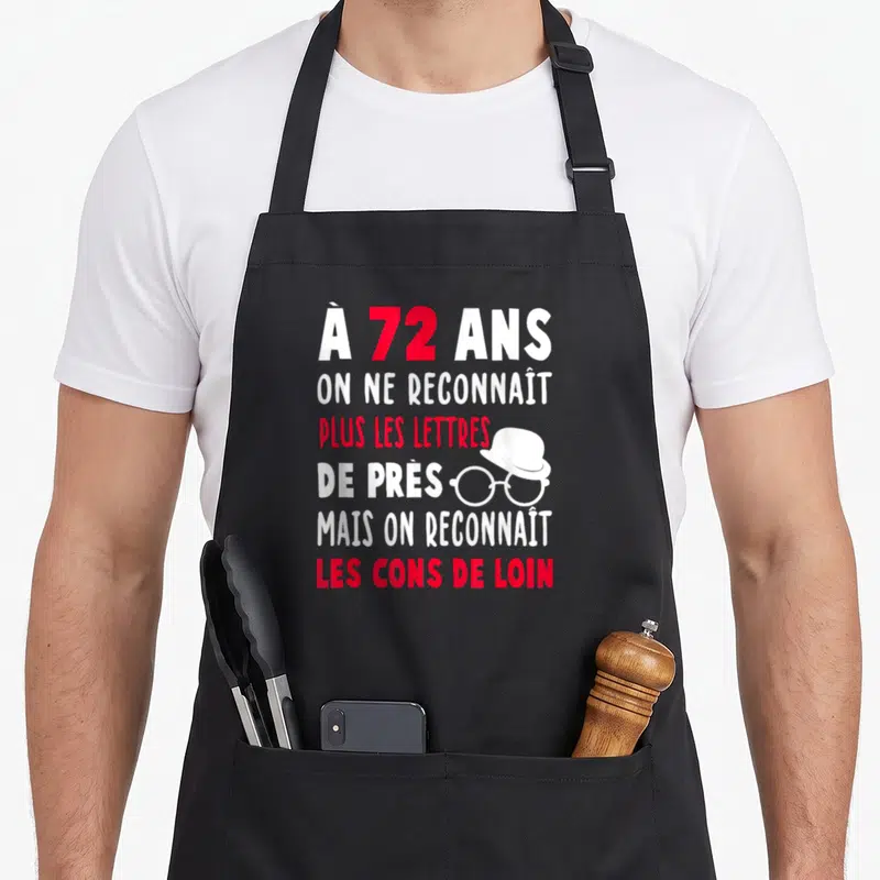 Tablier de cuisine noir avec texte blanc et rouge à 72 ans et illustration de lunettes et chapeau.