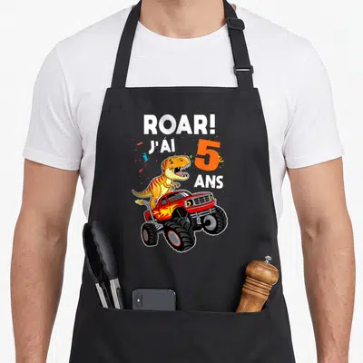 Tablier de cuisine noir avec un dinosaure sur un monster truck et l'inscription Roar J'ai 5 Ans entourée de confettis colorés.