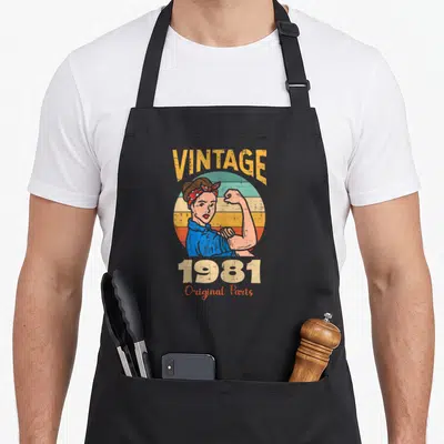 Tablier de cuisine noir avec illustration vintage Rosie Riveteuse et texte 1981 Original Parts en couleurs délavées.