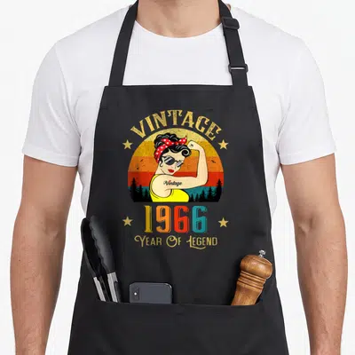 Tablier de cuisine noir avec illustration vintage d'une femme musclée en bandana rouge et lunettes de soleil sur fond arc-en-ciel.