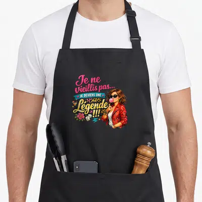 Tablier de cuisine noir avec une femme en veste rouge crachant du chewing-gum et le texte Je ne vieillis pas Je deviens une Légende