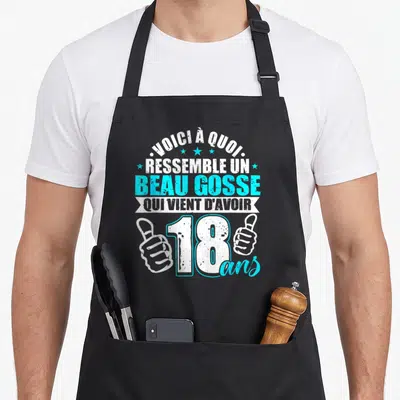 Tablier de cuisine noir avec inscription blanches et bleues célébrant les 18 ans d'un jeune homme, entouré de deux mains faisant des thumbs up.
