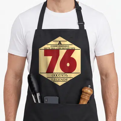 Tablier de cuisine noir avec vignette hexagonale beige affichant le chiffre 76 en rouge et texte humoristique sur fond uni.