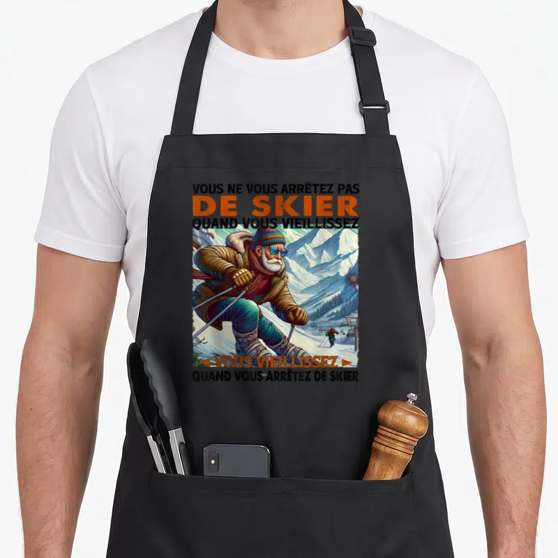 Tablier de cuisine noir avec illustration d'un skieur barbu portant des lunettes bleues et un bonnet coloré devant un paysage enneigé.
