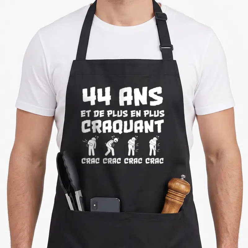 Tablier de cuisine noir avec texte blanc 44 ans et de plus en plus craquant et quatre personnages illustrés en train de se tordre.