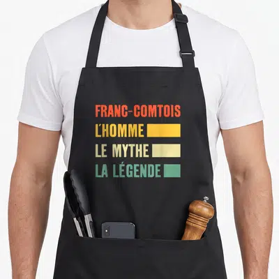 Tablier de cuisine noir avec inscription colorée Franc-Comtois LHomme Le Mythe La Légende en quatre lignes typographiques distinctes.