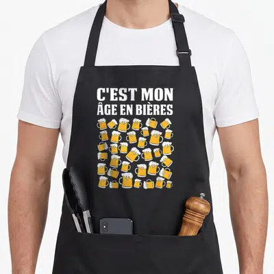 Tablier de cuisine noir avec l'inscription C'est mon âge en bières et des muglets de bière dorés à mousse blanche.