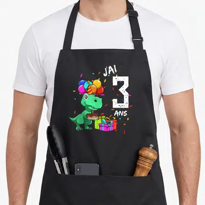 Tablier de cuisine noir avec un dinosaure vert tenant un gâteau et des cadeaux colorés.