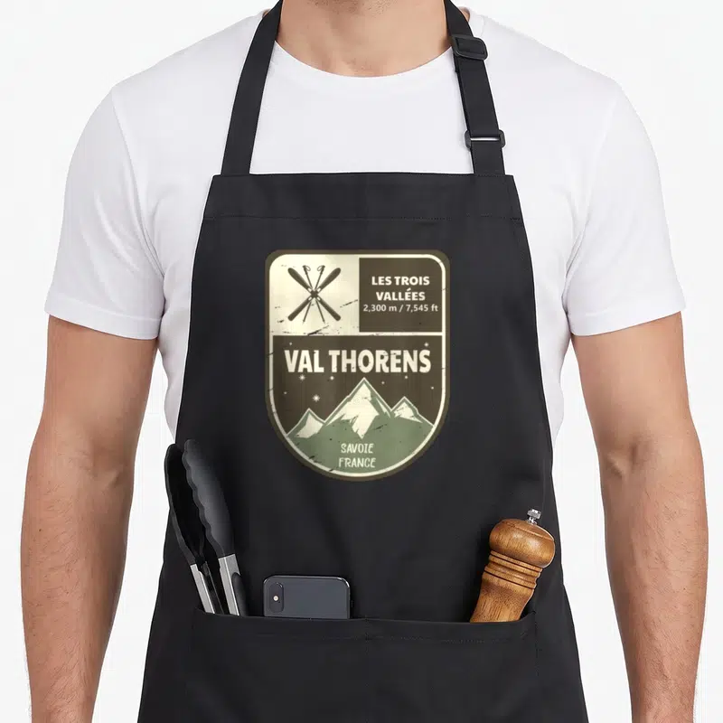 Tablier de cuisine noir avec un insigne vintage illustrant des skis croisés et les montagnes de Val Thorens en Savoie.