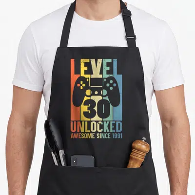 Tablier de cuisine noir avec illustration manette de jeu et texte Niveau 30 débloqué coloré sur fond strié.