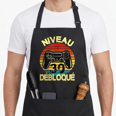 Tablier de cuisine noir avec illustration manette de jeu et texte Niveau 30 Débloqué sur fond de soleil retro.