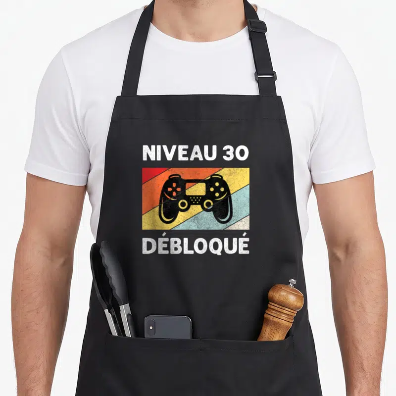 Tablier de cuisine noir avec manette de jeu et texte Niveau 30 Débloqué sur fond rayé multicolore.