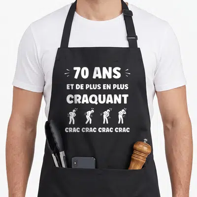 Tablier de cuisine noir avec inscription blanche 70 ans et de plus en plus craquant et quatre silhouettes stylisées.