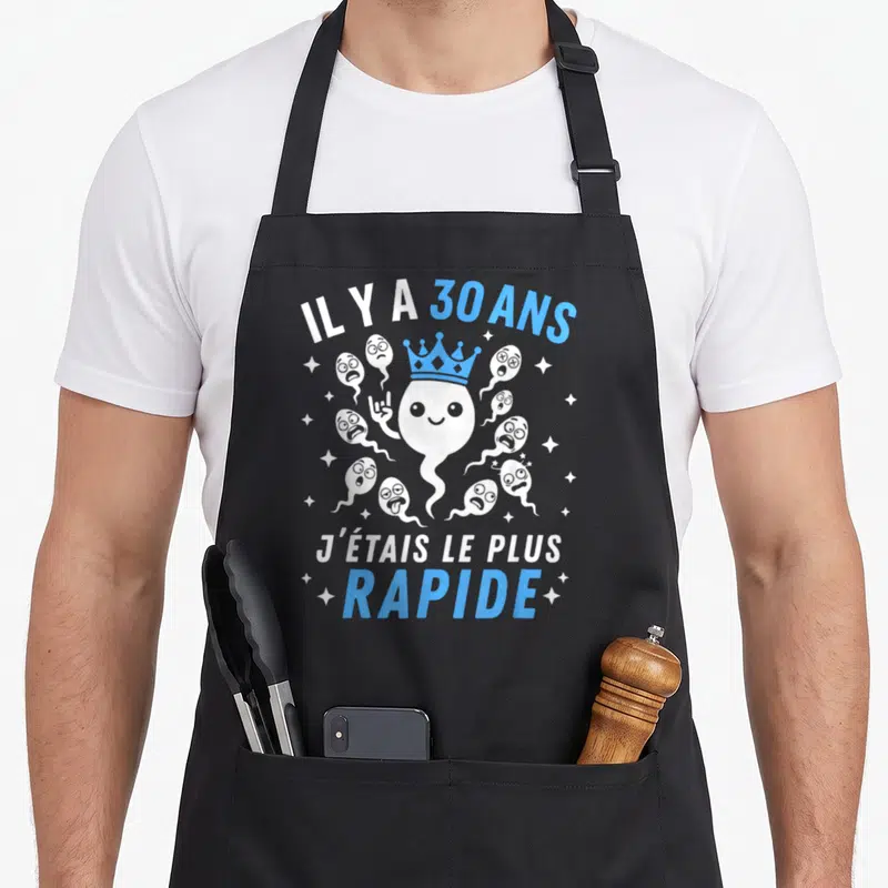 Tablier de cuisine noir avec illustration d'un spermatozoïde couronné et texte humoristique blanc et bleu.