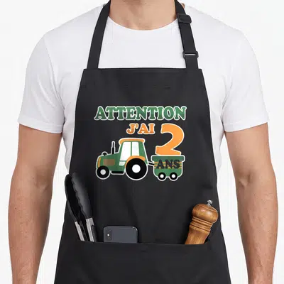 Tablier de cuisine noir avec illustration de tracteur vert et texte Attention j'ai 2 ans en lettres colorées.